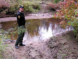 
DNR enforcement officer Jeff Bircham stands on an illegal trail that crosses the Warroad River. On the other side of the river, the DNR has felled trees to block the trail. Signs declaring the trail off limits are sometimes torn down by rogue riders. (MPR photo/Tom Robertson)